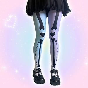 May include: Opaque tights featuring a split color design with a light blue left leg and a light purple right leg. The tights are printed with a black skeleton bone graphic that includes heart shapes at the knees and a row of stars along the side.