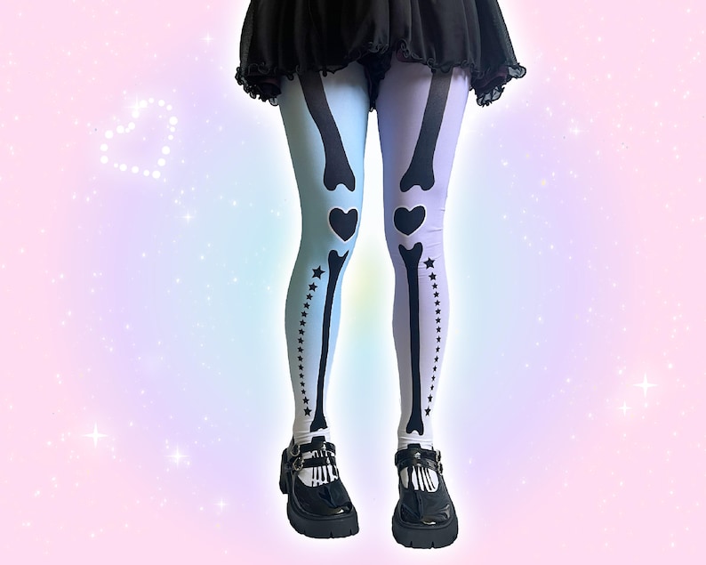 May include: Opaque tights featuring a split color design with a light blue left leg and a light purple right leg. The tights are printed with a black skeleton bone graphic that includes heart shapes at the knees and a row of stars along the side.