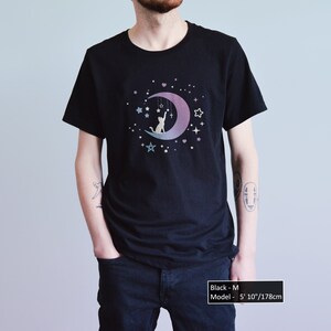 May include: Black t-shirt featuring a cat silhouette sitting on a crescent moon with stars and hanging decorations. The design has a gradient of purple and blue. The model is 5' 10"/178cm tall and is wearing a size M.