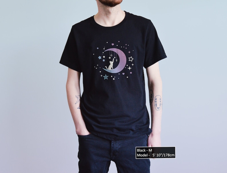 May include: Black t-shirt featuring a cat silhouette sitting on a crescent moon with stars and hanging decorations. The design has a gradient of purple and blue. The model is 5' 10"/178cm tall and is wearing a size M.