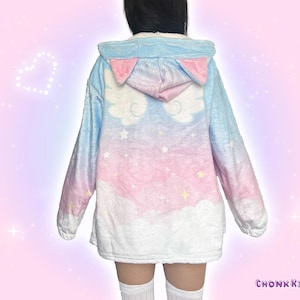 Könnte beinhalten: Eine Plüsch-Kapuzenjacke mit einem pastellfarbenen Farbverlauf in Blau, Rosa und Weiß mit Wolken- und Sternenmuster. Die Kapuze hat rosa Katzenohren und weiße Engelsflügel auf der Rückseite. Der Text CHONK KITTEA ist unten aufgedruckt.