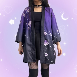 May include: A kimono-style jacket in shades of purple and black, adorned with white floral patterns. The jacket has a gradient effect, transitioning from a deep purple at the top to black at the bottom. The sleeves are wide, and the fabric appears to be silky.