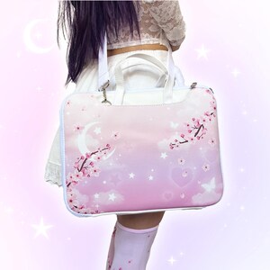 May include: A pink laptop bag with a dreamy design featuring a crescent moon, cherry blossoms, stars, and hearts. The bag has a white handle and strap, and a white border. The background is a gradient of pink and purple.