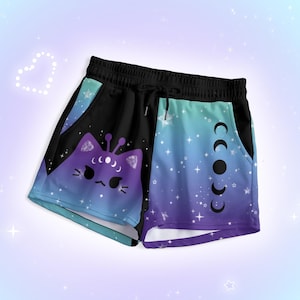 Op de afbeelding: Zwarte shorts met trekkoord en een ontwerp met kattengezicht en maanfasen. De shorts hebben een verloop van blauw, paars en zwart, met een kattengezicht aan de ene kant en maanfasen aan de andere kant. Comfortabele pasvorm.