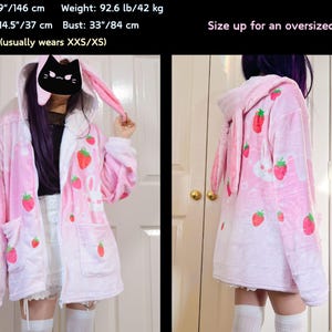 Puede incluir: Sudadera de felpa rosa con capucha de orejas de conejo y estampado de fresas y conejos. La sudadera tiene cierre de cremallera y dos bolsillos. La modelo lleva una talla S y mide 1,46 m de altura y pesa 42 kg. El texto "Size up for an oversized look!" es visible en la imagen.
