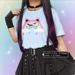 K&ouml;nnte beinhalten: Hellblaues Unisex-T-Shirt mit einem Regenbogen-Katzen-Design, mit Flederm&auml;usen und tropfenden Wolken. Das Model tr&auml;gt einen schwarzen Rock und Armstulpen. Der Text auf dem Shirt lautet "Heather Ice Blue - M (Unisex)" und das Model ist 146cm gro&szlig;.