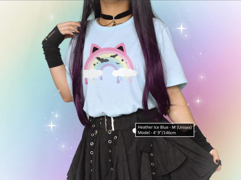 K&ouml;nnte beinhalten: Hellblaues Unisex-T-Shirt mit einem Regenbogen-Katzen-Design, mit Flederm&auml;usen und tropfenden Wolken. Das Model tr&auml;gt einen schwarzen Rock und Armstulpen. Der Text auf dem Shirt lautet "Heather Ice Blue - M (Unisex)" und das Model ist 146cm gro&szlig;.