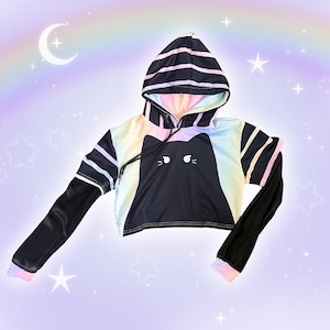 May include: Black cropped hoodie with a cat face design. The hoodie features a rainbow gradient on the front and hood, with black sleeves and striped accents. The background is a pastel purple with a rainbow, moon, and stars.