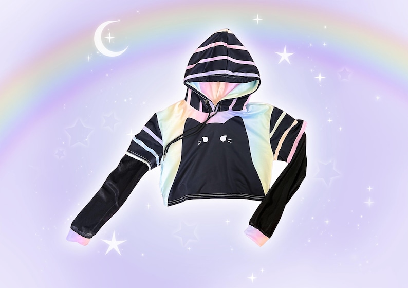 May include: Black cropped hoodie with a cat face design. The hoodie features a rainbow gradient on the front and hood, with black sleeves and striped accents. The background is a pastel purple with a rainbow, moon, and stars.