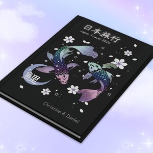 May include: A black journal with a Japanese design featuring two koi fish, cherry blossoms, and the text "日本旅行 Japan Travel 2025". The journal also has the text "Christine & Daniel" on the front cover.