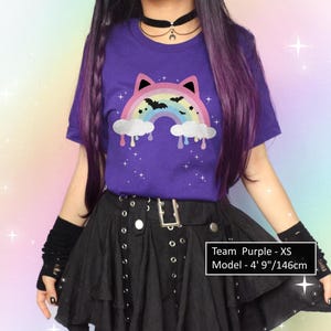 K&ouml;nnte beinhalten: Ein lila T-Shirt mit einem Regenbogen- und Fledermaus-Motiv. Der Regenbogen hat Katzenohren und tropfende Wolken. Die Person tr&auml;gt einen schwarzen Rock und fingerlose Handschuhe. Der Text auf dem Bild lautet: "Team Purple - XS Model - 146cm."