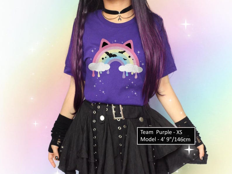 K&ouml;nnte beinhalten: Ein lila T-Shirt mit einem Regenbogen- und Fledermaus-Motiv. Der Regenbogen hat Katzenohren und tropfende Wolken. Die Person tr&auml;gt einen schwarzen Rock und fingerlose Handschuhe. Der Text auf dem Bild lautet: "Team Purple - XS Model - 146cm."