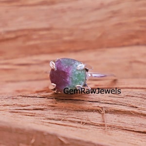 May include: A silver ring featuring a raw, uncut gemstone with a mix of red and green hues. The gemstone is held in place by four prongs. The ring is set against a wooden background, with the text "GemRawJewels" visible.