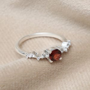 Può includere: Un anello d'argento con una gemma rotonda di colore rosso intenso al centro, affiancata da pietre chiare più piccole. L'anello ha un design delicato e curvo, su uno sfondo neutro e strutturato. Un gioiello.