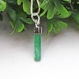 May include: A green cylindrical pendant hangs from a silver chain. The pendant has a silver cap and a bright green, textured body. The necklace is displayed against a backdrop of green leaves and a white surface.
