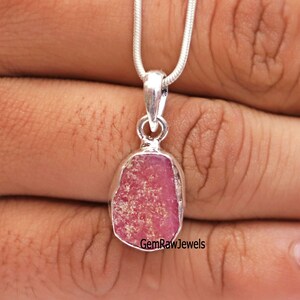 Puede incluir: Un collar con colgante de plata con una piedra preciosa rosa en bruto. El colgante tiene una forma irregular y está engastado en un bisel de plata. La cadena del collar es una fina cadena de serpiente plateada. El texto "GemRawJewels" es visible en la parte inferior derecha.