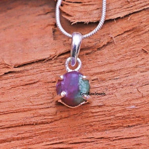 Peut inclure: Un collier pendentif argenté orné d'une pierre précieuse brute multicolore. La pierre affiche un mélange de teintes violettes profondes et vertes, sertie dans un métal doré. Le collier est présenté sur une surface en bois.