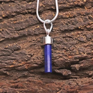 Może przedstawiać: Srebrny naszyjnik z wisiorkiem z ciemnoniebieskiego, sześciokątnego kamienia szlachetnego. Cylindryczny kamień szlachetny ma srebrną nasadkę. Naszyjnik jest prezentowany na tle teksturowanego, brązowego drewnianego tła.