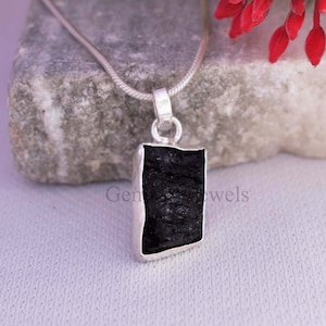 May include: A silver pendant necklace featuring a rectangular, raw-cut black gemstone. The stone is set in a silver bezel and hangs from a delicate silver chain. The background includes a gray stone and red flowers.