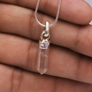 May include: A clear quartz crystal pendant with a silver-colored metal cap and bail, suspended from a thin, silver-colored chain. The crystal is a light, translucent color and is shaped into a pointed, elongated form. The pendant is a simple, elegant piece of jewelry.