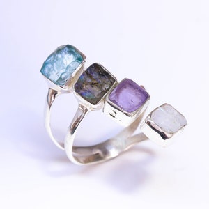 Può includere: Un anello d'argento con quattro pietre quadrate nei toni del blu, viola e bianco. Le pietre sono incastonate in castoni individuali e disposte in fila sulla parte superiore dell'anello.