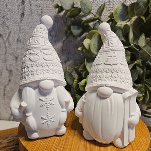 Könnte beinhalten: Zwei weiße Keramik-Gnomfiguren mit winterlichen Hüten und gemusterten Details. Ein Gnom hält einen Skistock und der andere hat ein Schneeflockenmuster auf der Brust.
