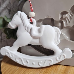 Könnte beinhalten: Weißer Keramik-Schaukelpferdornament mit einer Schnur und einer Perle zum Aufhängen. Das Ornament befindet sich in einer Silikonform.