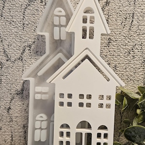 Könnte beinhalten: Weiße hausförmige Formen mit Fensterausschnitten. Die Formen befinden sich vor einem strukturierten grauen Hintergrund. Die Hausformen haben ein schlichtes, klares Design, perfekt für Bastelprojekte.