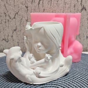 Könnte beinhalten: Weißer Keramik-Pflanztopf in Form eines Mädchens mit langen Haaren, das ein kleines Tier hält. Der Topf steht neben einer rosa Silikonform.