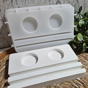 Könnte beinhalten: Weiße Silikonform zum Herstellen von zweistufigen Kerzenhaltern. Die Form hat zwei runde Vertiefungen für die Kerzenhalter und eine rechteckige Vertiefung für die Basis. Die Form ist aus flexiblem Silikon und einfach zu bedienen.