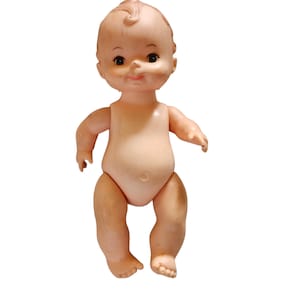 May include: A vintage baby doll with a light skin tone. The doll has a round face, dark eyes, and a small, smiling mouth. Its arms and legs are slightly bent, and it has a small belly button. The doll's hair is styled in a small curl on top of its head.