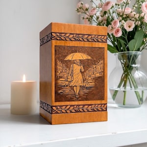 Handcrafted wooden cremation urn box featuring a detailed engraved rainy street scene with a woman walking under an umbrella. This memorial urn is made from polished natural wood with decorative carved leaf borders. A beautiful funeral urn designed for safely holding ashes while serving as an elegant remembrance keepsake and memorial tribute for a loved one.