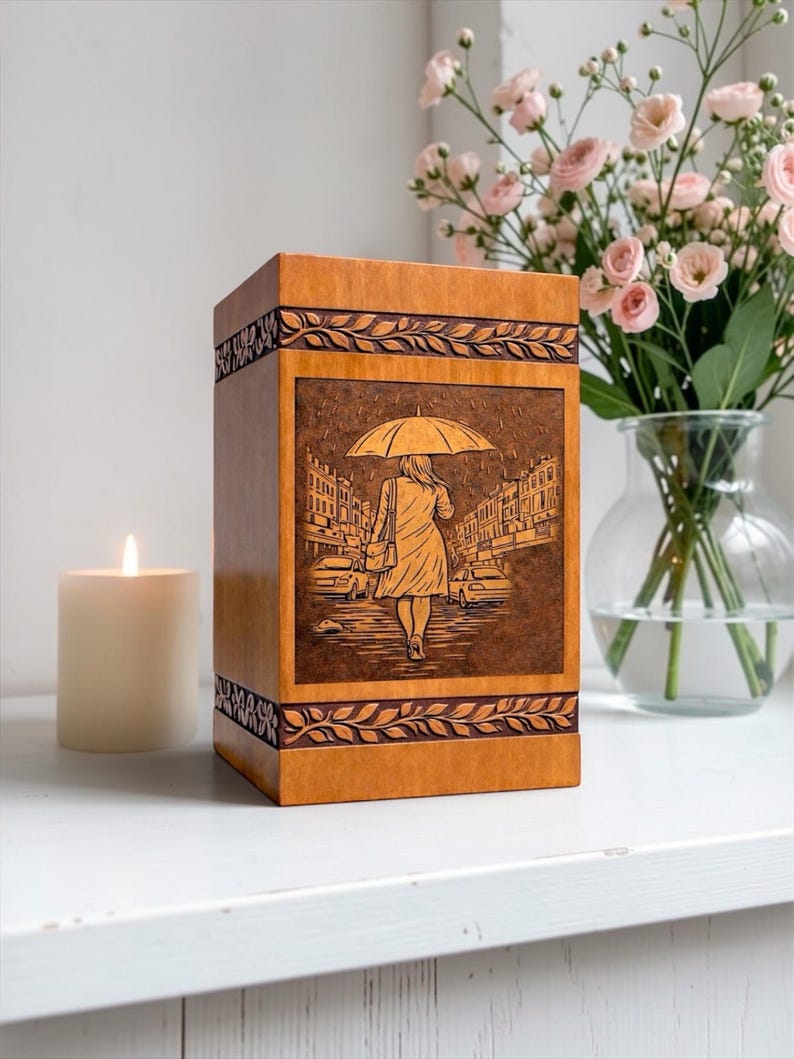 Handcrafted wooden cremation urn box featuring a detailed engraved rainy street scene with a woman walking under an umbrella. This memorial urn is made from polished natural wood with decorative carved leaf borders. A beautiful funeral urn designed for safely holding ashes while serving as an elegant remembrance keepsake and memorial tribute for a loved one.