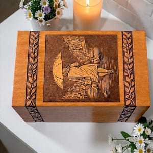 Handmade wooden urn for ashes featuring artistic rain walk engraving and detailed city street background. The urn box is crafted from durable wood with ornamental carved leaf borders and a warm finish. This memorial keepsake urn is ideal for honoring and remembering a loved one with dignity and beauty.