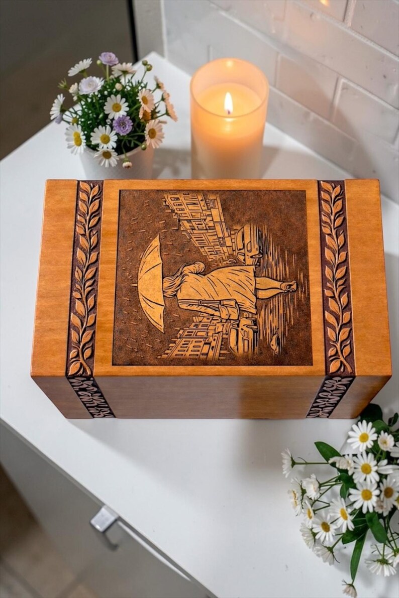 Handmade wooden urn for ashes featuring artistic rain walk engraving and detailed city street background. The urn box is crafted from durable wood with ornamental carved leaf borders and a warm finish. This memorial keepsake urn is ideal for honoring and remembering a loved one with dignity and beauty.
