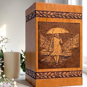 Premium wooden cremation urn box with engraved memorial artwork of a woman walking under an umbrella in the rain. Crafted from high-quality natural wood with decorative carved borders, this funeral urn provides a respectful and beautiful resting place for ashes while serving as a timeless tribute to a loved one's memory.