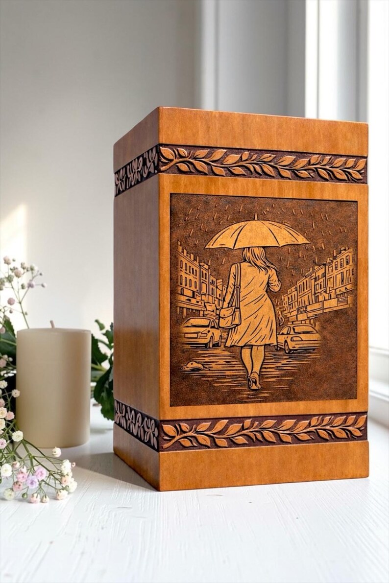Premium wooden cremation urn box with engraved memorial artwork of a woman walking under an umbrella in the rain. Crafted from high-quality natural wood with decorative carved borders, this funeral urn provides a respectful and beautiful resting place for ashes while serving as a timeless tribute to a loved one's memory.