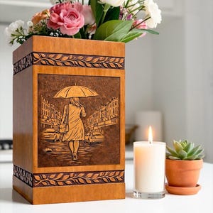 Elegant handcrafted wooden urn box for ashes with a unique umbrella rain walk engraving design. The detailed artwork shows a peaceful figure walking along a city street, symbolizing remembrance and life's journey. This wooden cremation urn includes carved border accents and a warm natural wood finish, making it a meaningful memorial keepsake.