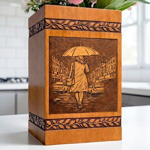Decorative wooden memorial urn box with intricate engraving of a rainy street scene and umbrella figure. The handcrafted design features elegant leaf borders and a smooth polished wood finish. This cremation urn for human ashes is designed for safe storage while creating a peaceful and meaningful remembrance display in the home.