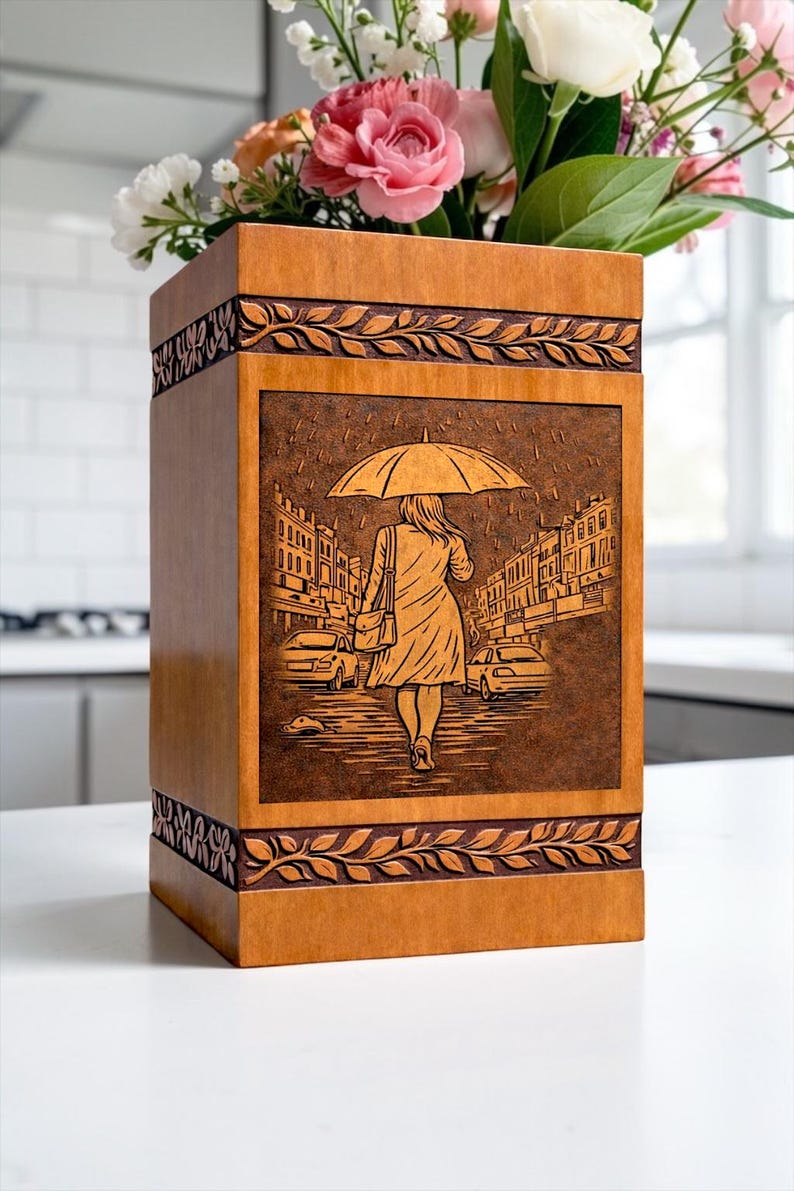 Decorative wooden memorial urn box with intricate engraving of a rainy street scene and umbrella figure. The handcrafted design features elegant leaf borders and a smooth polished wood finish. This cremation urn for human ashes is designed for safe storage while creating a peaceful and meaningful remembrance display in the home.