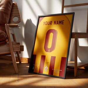 May include: A framed print of a yellow and red soccer jersey with the number 0 on the front. The jersey has a black frame and the text "YOUR NAME" at the top. The jersey also has a small logo on the bottom left corner.