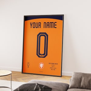 Op de afbeelding: Een ingelijste poster met een oranje achtergrond en een donkerblauw nummer "0" in het midden. De poster heeft de tekst "YOUR NAME" bovenaan en "NEDERLANDS ELFTAL" onderaan. De poster is ingelijst in zwart.