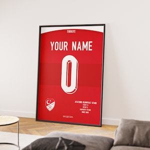Op de afbeelding: Een rode en witte poster met de tekst "YOUR NAME" en het nummer "0" in een witte cirkel. De poster bevat ook een voetbalteamlogo en de tekst "TURKIYE" en "ATATURK OLYMPIAT STAD".