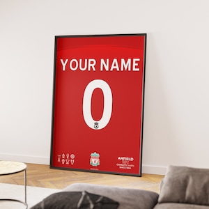 Peut inclure: Affiche encadrée d'un maillot de sport rouge avec le texte "YOUR NAME" au-dessus d'un grand "0" blanc. Le maillot présente l'écusson du Liverpool FC et le mot "ANFIELD". Le cadre est noir.