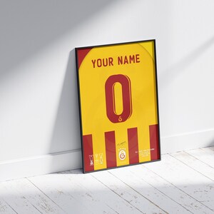 May include: A framed print of a yellow and red soccer jersey with the number "0" in red and the text "YOUR NAME" at the top. The jersey has a red and yellow striped pattern and a signature at the bottom.