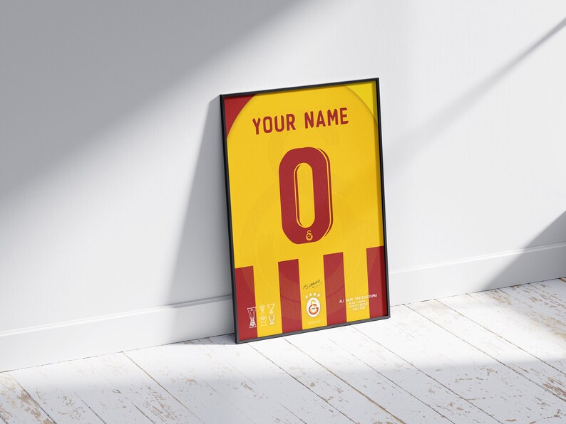 May include: A framed print of a yellow and red soccer jersey with the number "0" in red and the text "YOUR NAME" at the top. The jersey has a red and yellow striped pattern and a signature at the bottom.