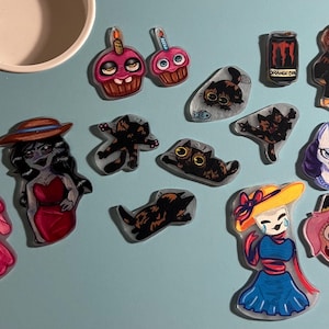 May include: A collection of assorted acrylic charms featuring various illustrated characters, cats, cupcakes, and a beverage can. The designs include a pink-haired figure, a character in a red dress, a blue-haired unicorn, and a pink-haired child character.