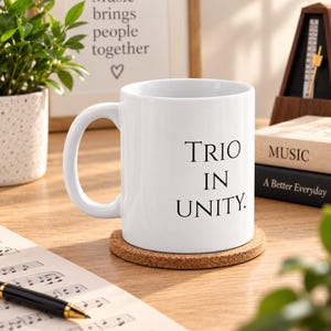 May include: A white ceramic mug with a handle, featuring the black printed text "TRIO IN UNITY." The mug sits on a round, light brown cork coaster. The design is simple, with a smooth finish and a standard cylindrical shape for holding beverages.