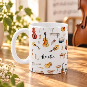 Op de afbeelding: Een witte keramische mok met een herhalend aquarelpatroon van diverse snaarinstrumenten, waaronder violen en cello's, samen met muzieknotatie. De naam Amelia staat in een zwart cursief lettertype op de voorzijde gedrukt.