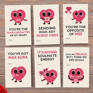 May include: A collection of six Valentine's Day cards with heart-shaped characters and playful messages. The cards feature phrases like "You're the main character of my heart" and "It's giving soulmate energy." The cards are on a wooden surface.